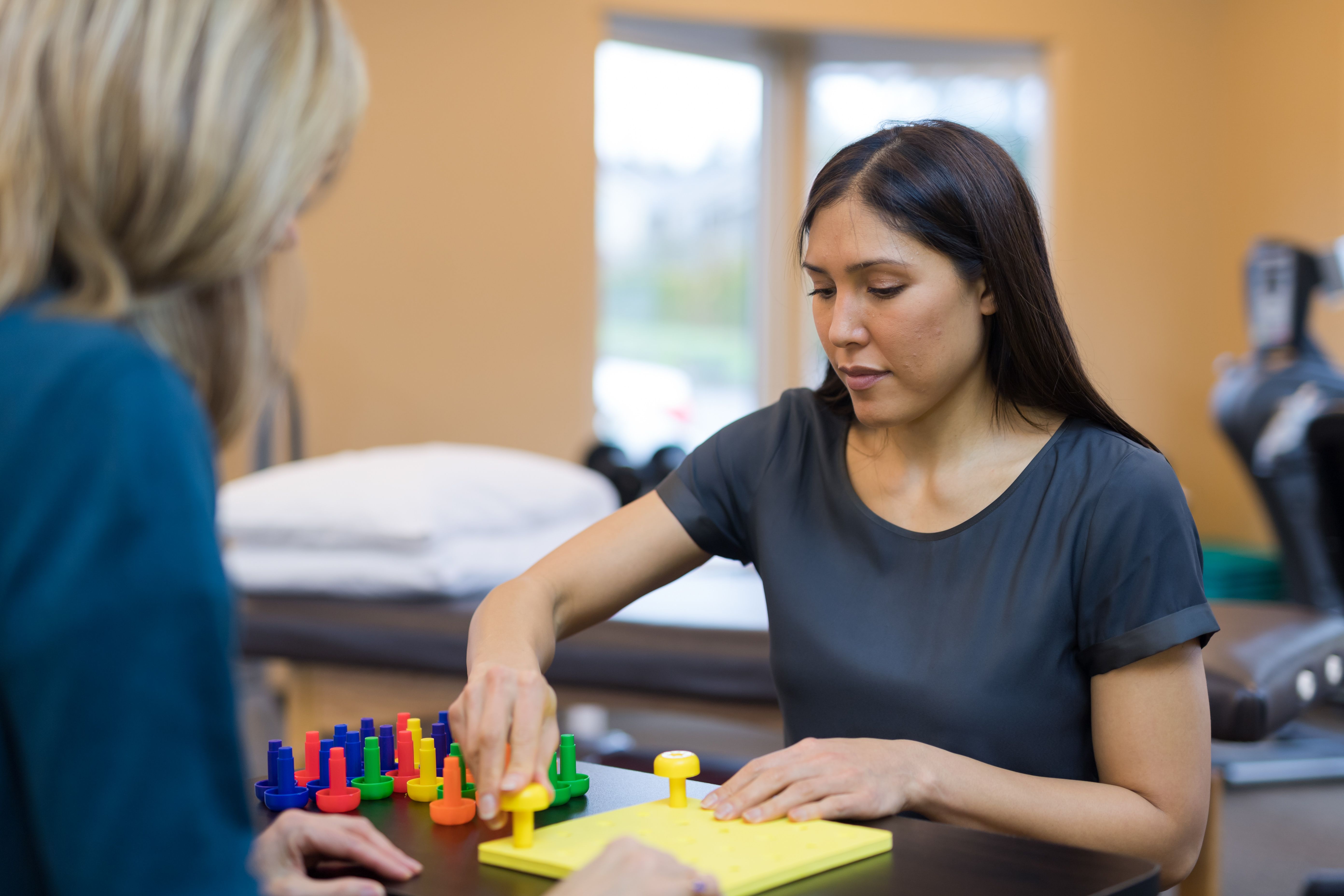An occupational therapist works with a woman on her coordination and motor skills. They are doing a fun exercise putting pegs into a plastic board. The shot is over the patient's shoulder and is focused on the therapist.